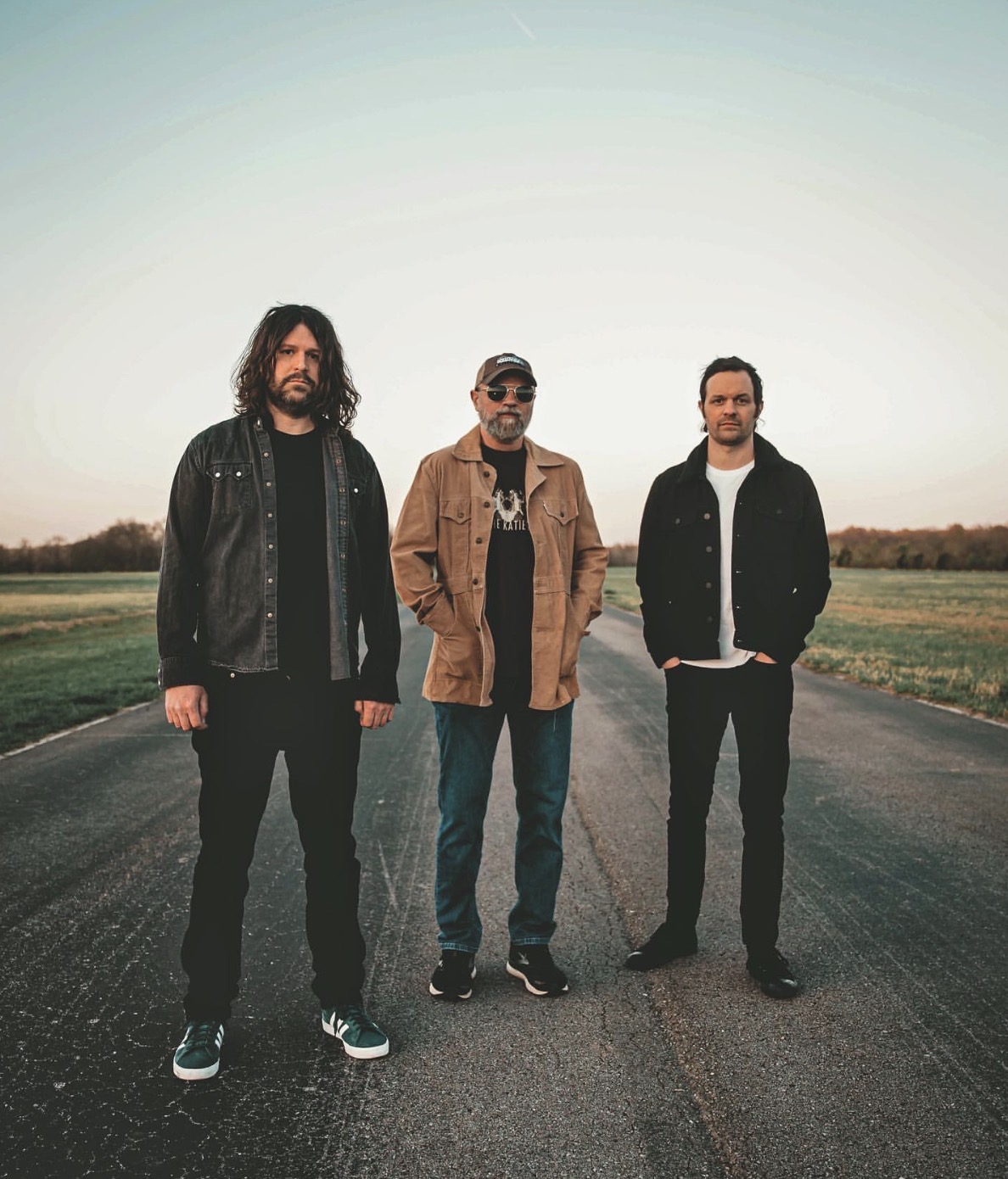 Three men stand apart on a deserted rural road at sunset, looking at the camera. They wear casual jackets and jeans, with the middle man in a tan jacket and cap.